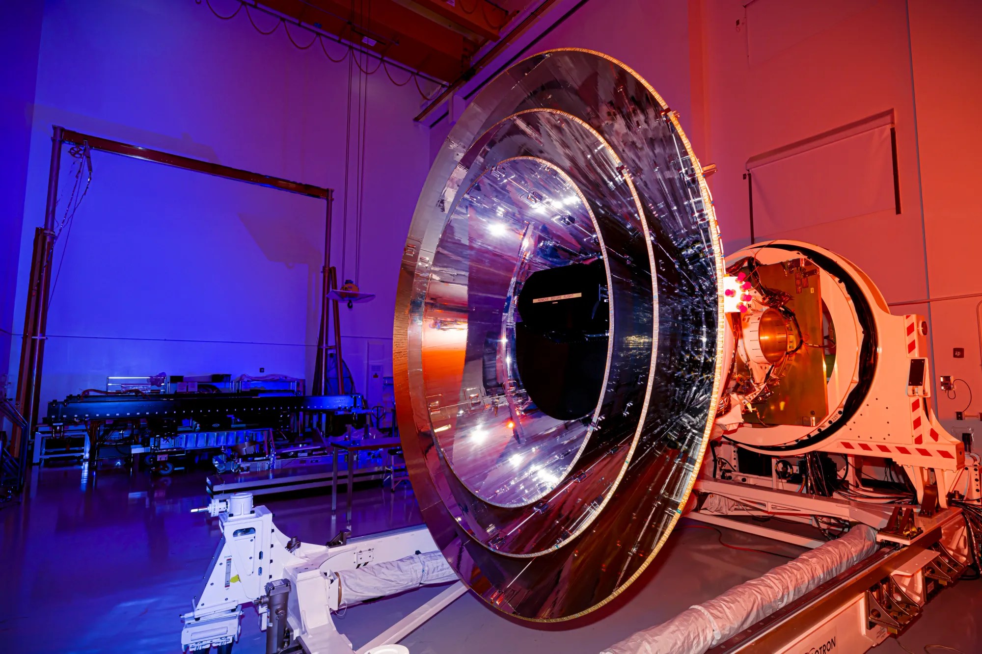 The large, shiny silver, round SPHEREx telescope sits in a room lit by blue and orange lighting.