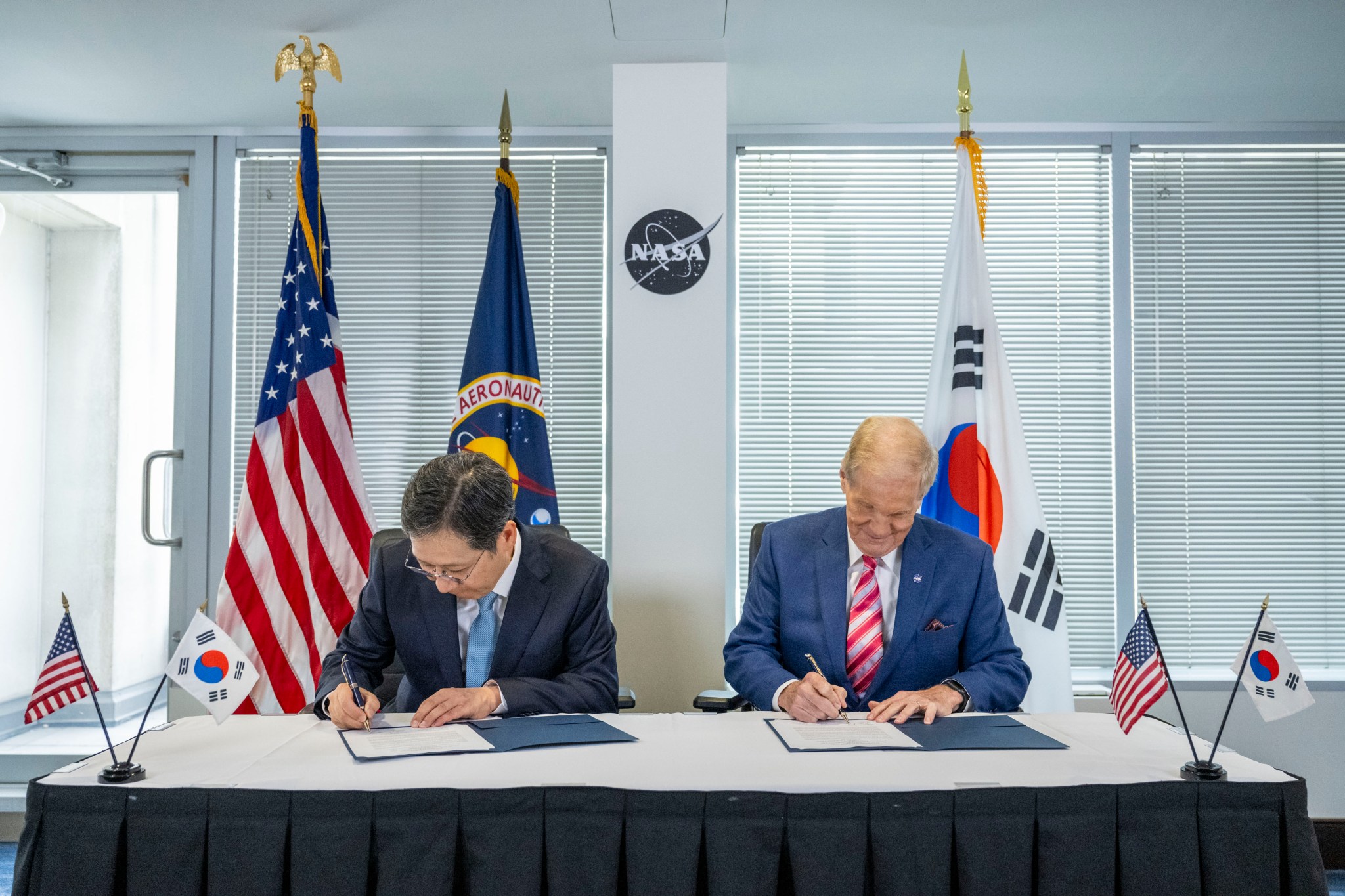 Korea AeroSpace Administration (KASA) Administrator Youngbin Yoon, left, and NASA Administrator Bill Nelson, right, sit at a table at NASA Headquarters signing a document with flags of each country behind them.