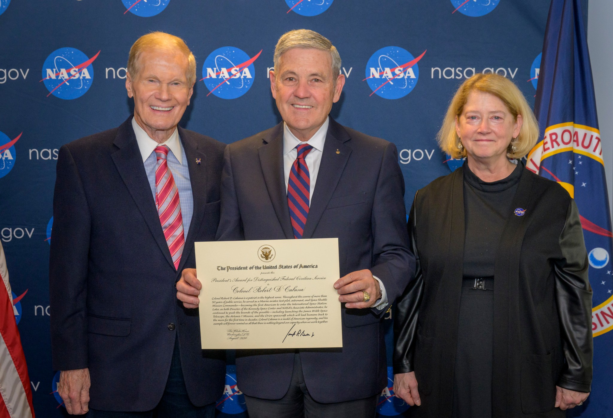 Nelson, Cabana, and Melroy are side by side, smiling, in front of a background with NASA logos, with Cabana holding the award certificate.