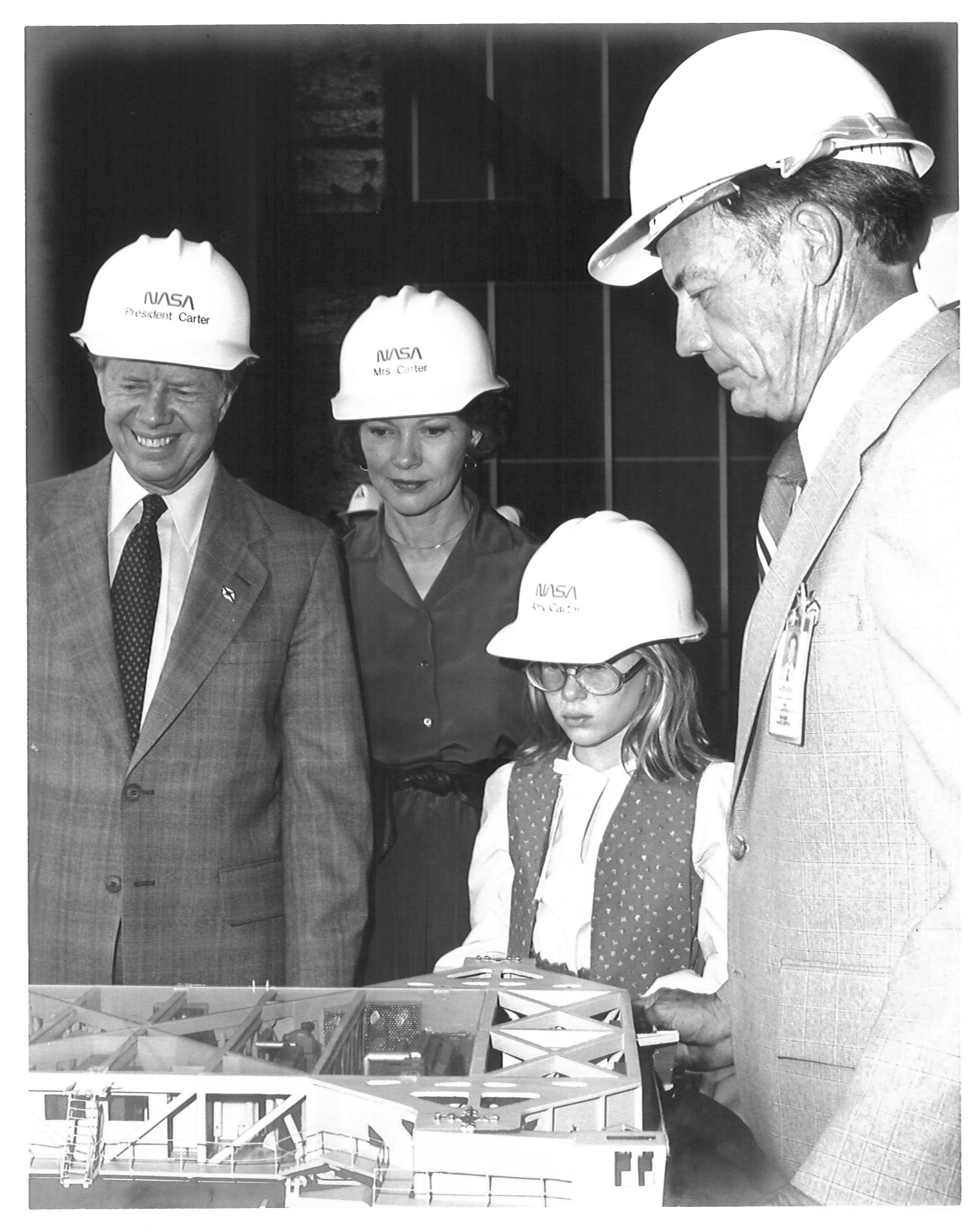 President Jimmy Carter, wife Rosalynn, daughter Amy, and Kennedy Space Center director Lee Scherer, all wearing NASA hard hats, look down at a scale model of the crawler, which transported the Shuttle to the launch pad. The photo is in black and white.