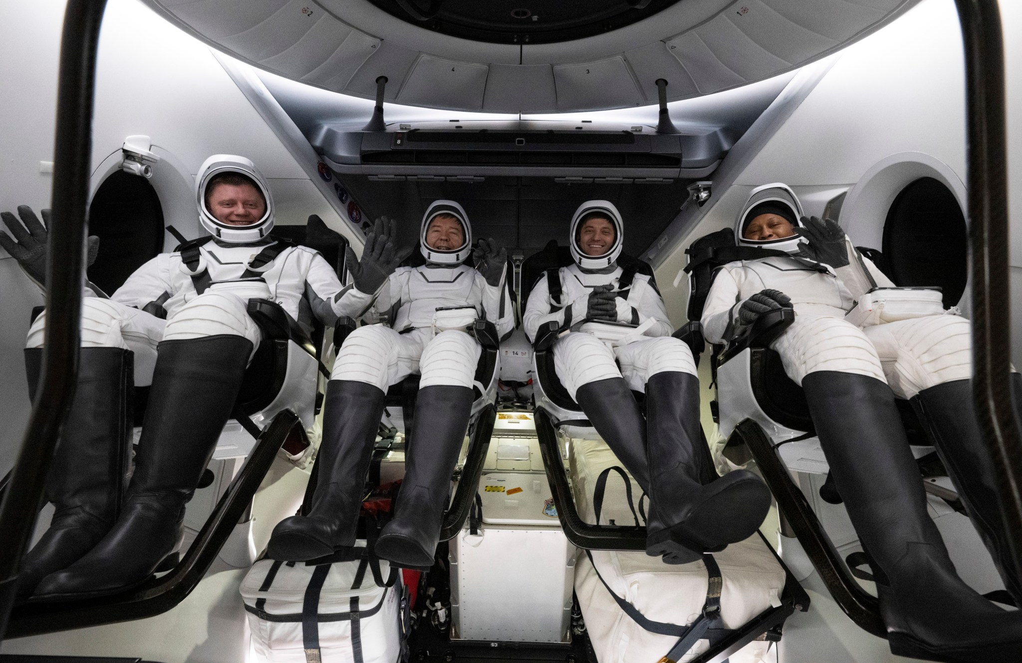 The four NASA SpaceX Crew-8 members sit in a row, smiling and waving in their spacesuits aboard a Dragon spacecraft.