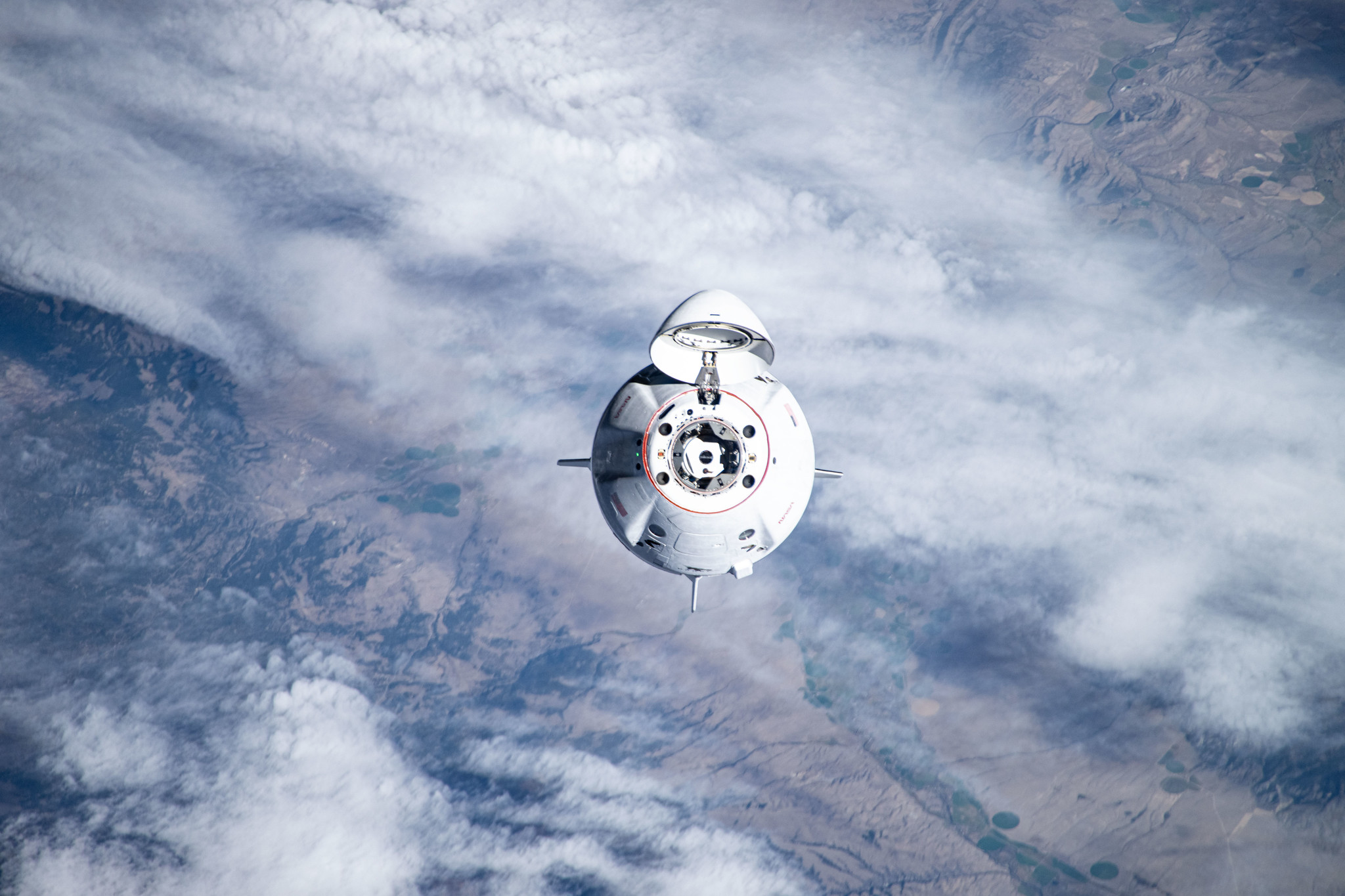 A white SpaceX Dragon spacecraft, with its nose cone open, is seen above Earth's blue water and white clouds.