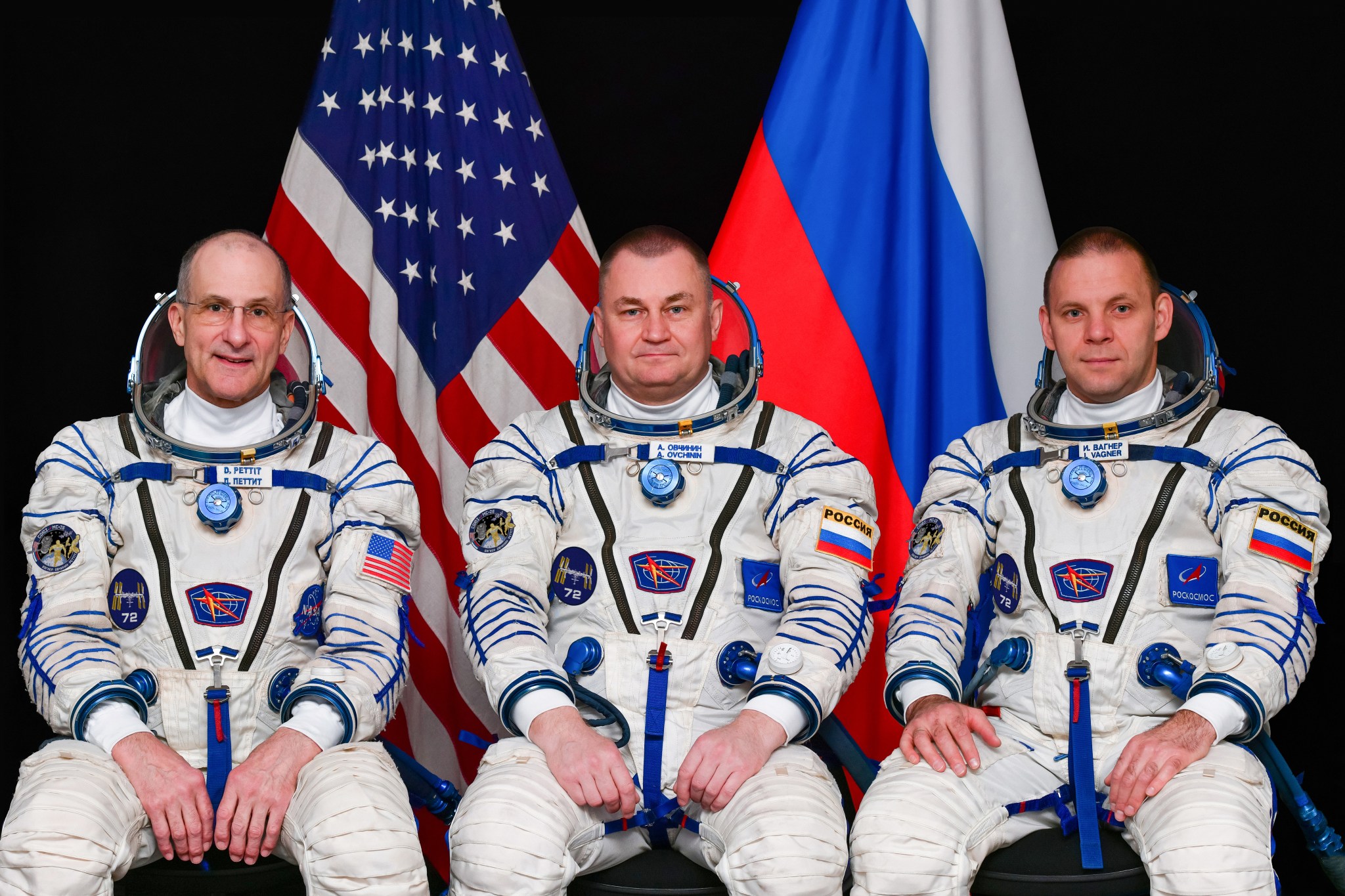 A portrait image of a NASA astronaut and two Roscosmos cosmonauts sitting in front of the flags of the United States and Russia against a black backdrop.